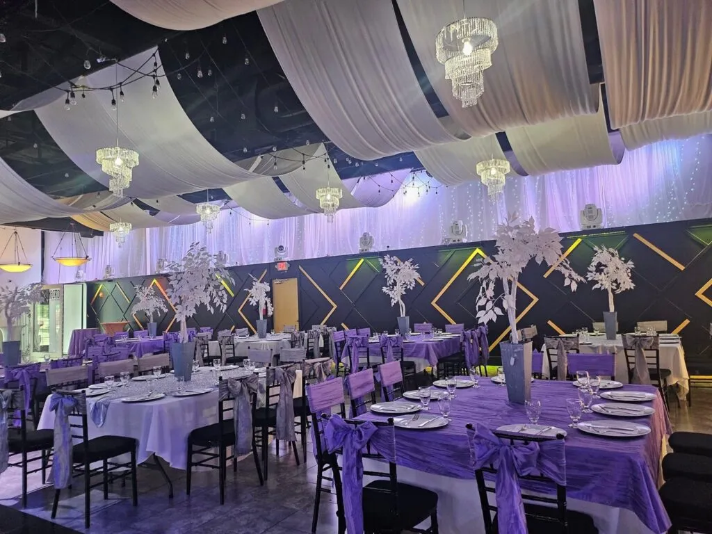 Banquet tables in lavender and white beneath a draped ceiling and chandeliers.